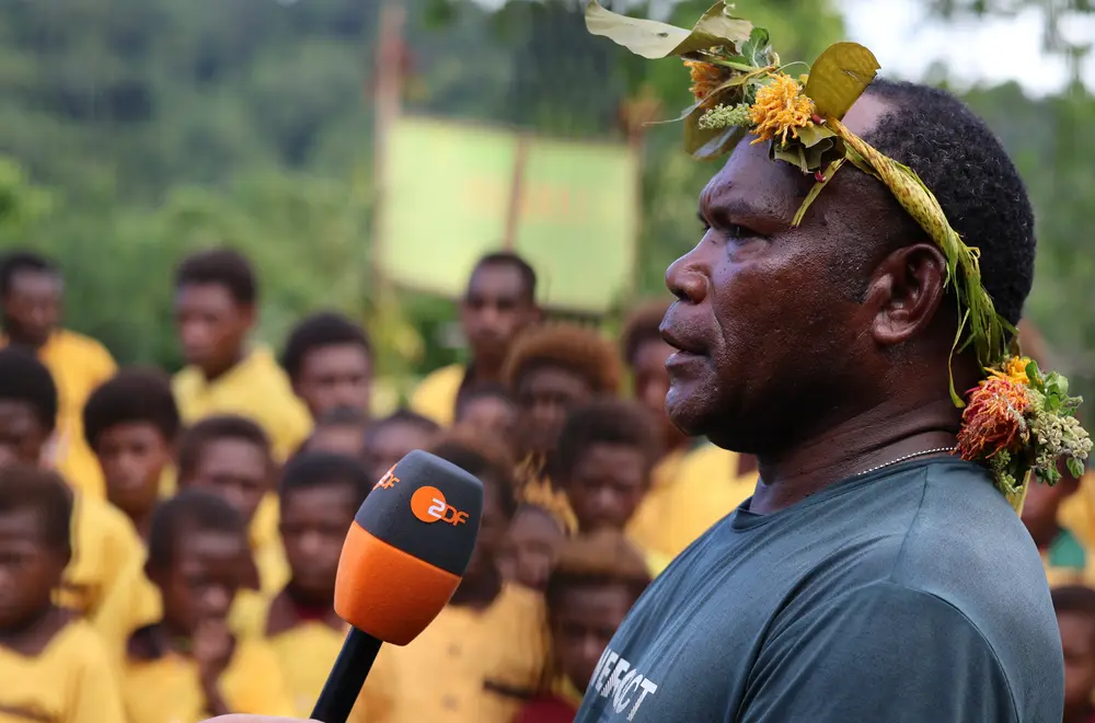 Peter Kikele spricht in ein ZDF-Mikrofon, Personen im Hintergrund.