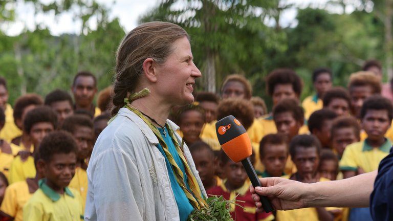 Eine Frau mit Naturschmuck spricht ins ZDF-Mikrofon, hinten eine Schulklasse. 